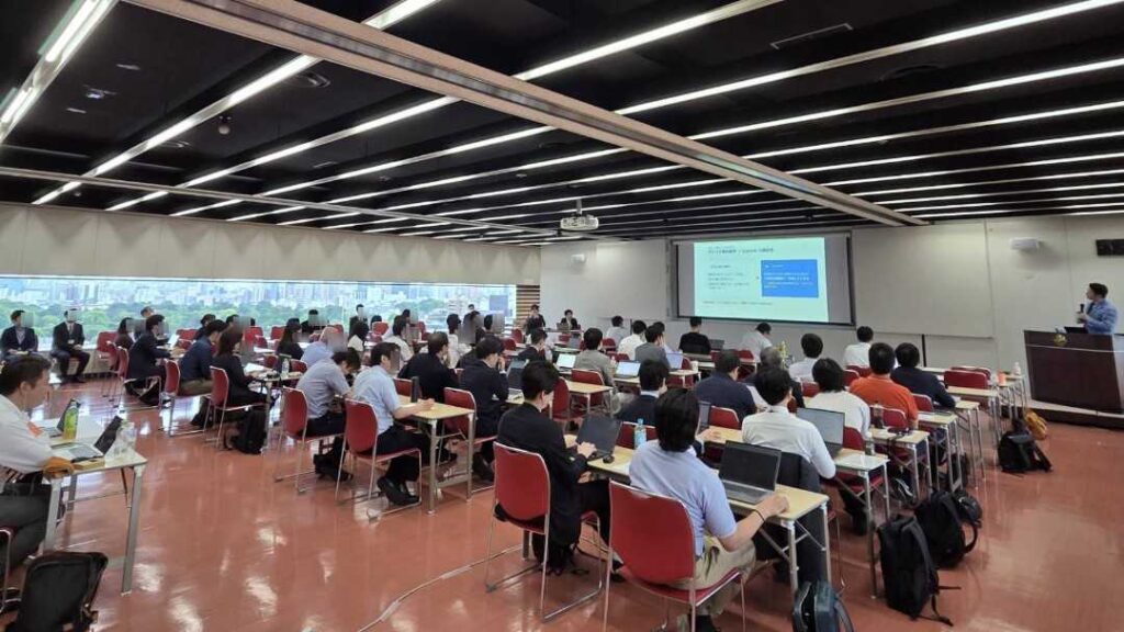 Large lecture hall with rows of red chairs, attendees using laptops, and a presenter at a podium by a projector screen at the front.