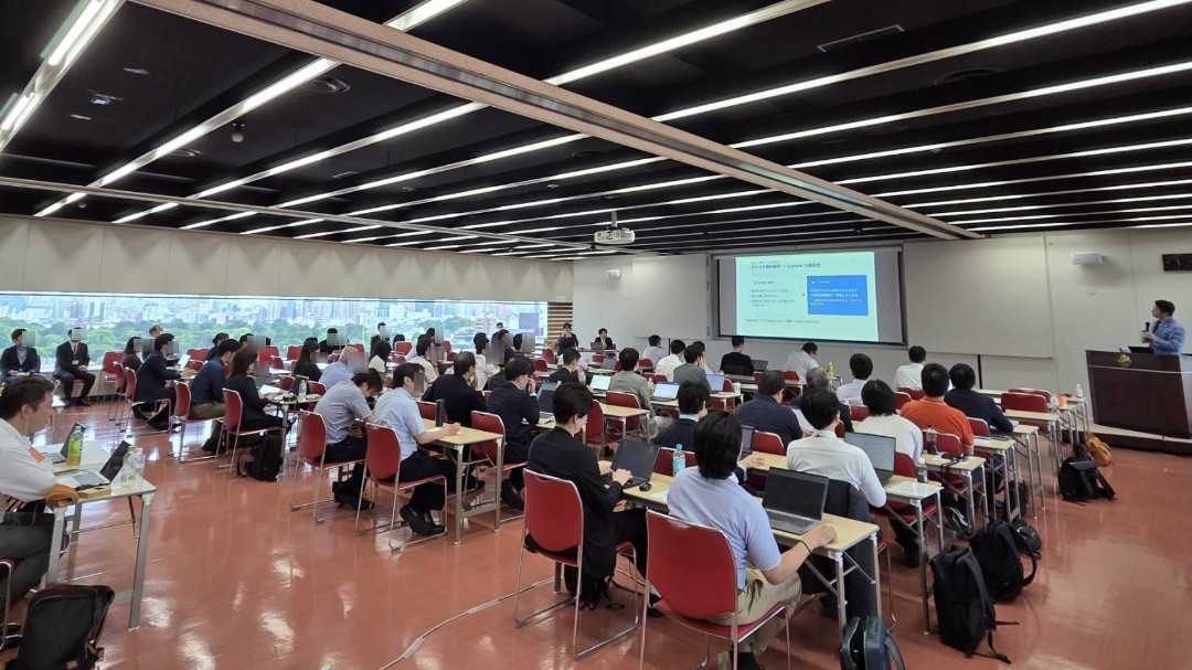 Large lecture hall with rows of red chairs, attendees using laptops, and a presenter at a podium by a projector screen at the front.